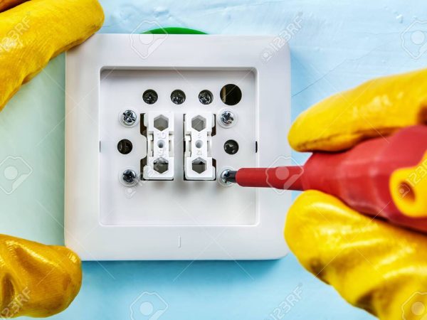 Worker in yellow protective gloves is tightening screw in european standard light switch with dielectric screwdriver.